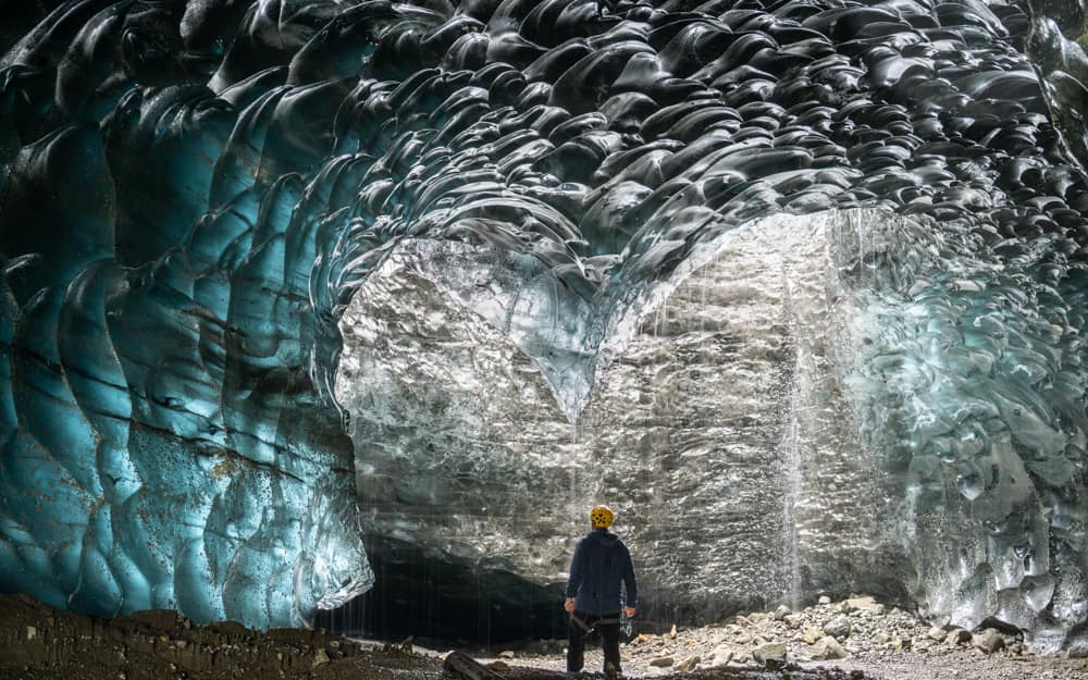 A person in a yellow helmet stands under the rippled blue and clear ice of a natural cave in Vatnajökull